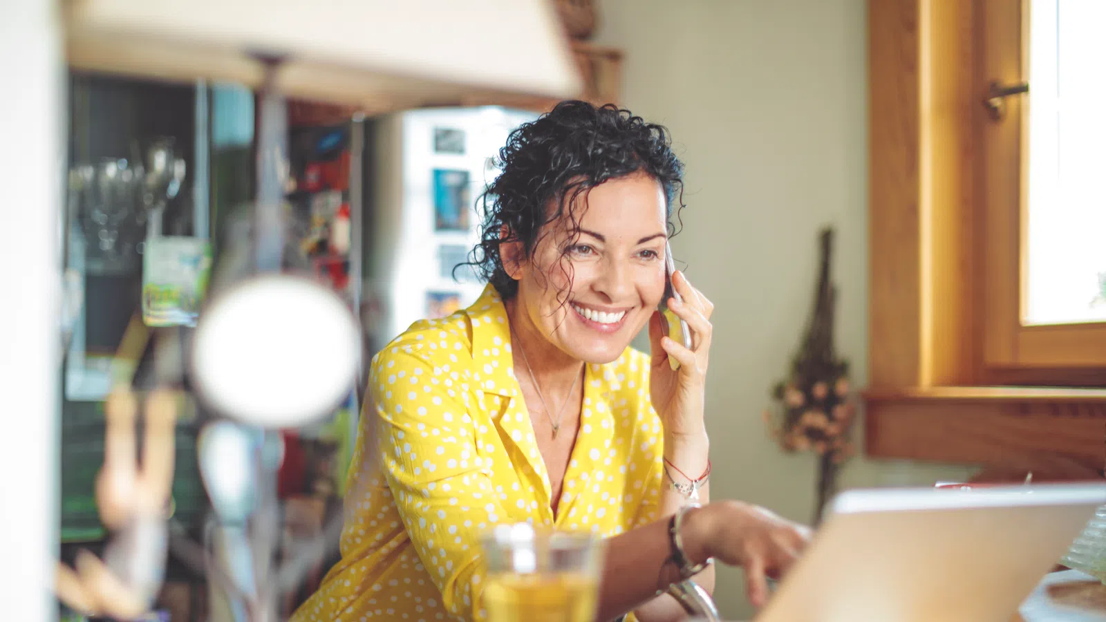 Smiling woman in yellow shirt multitasking with phone and laptop at kitchen table with drink and food.