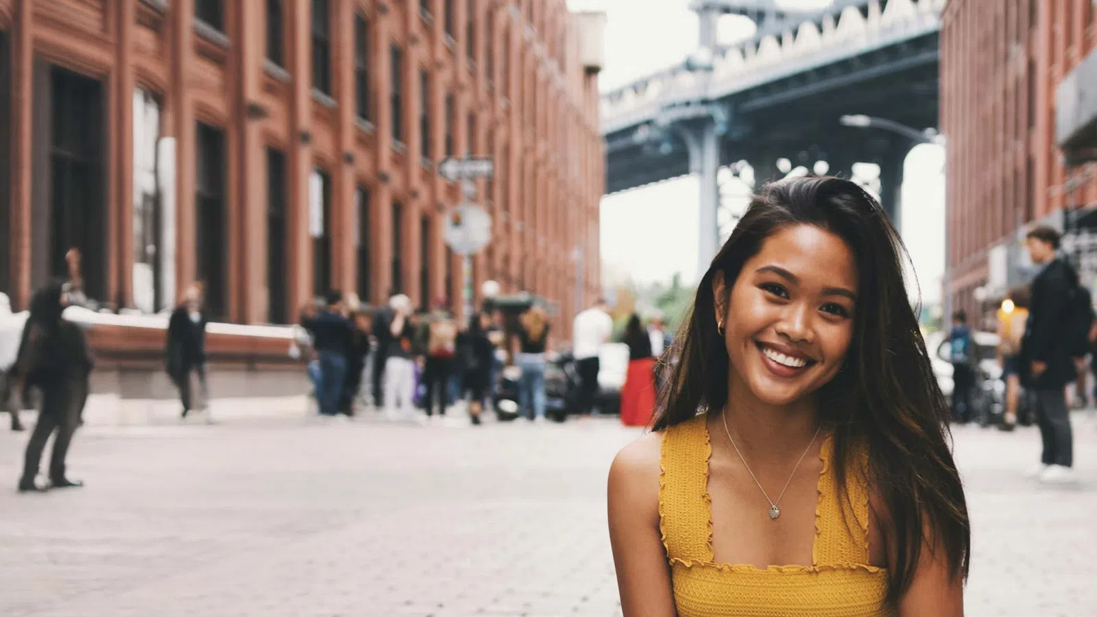 Smiling young woman in a yellow top sitting on a cobblestone street with brick buildings and a bridge in the background