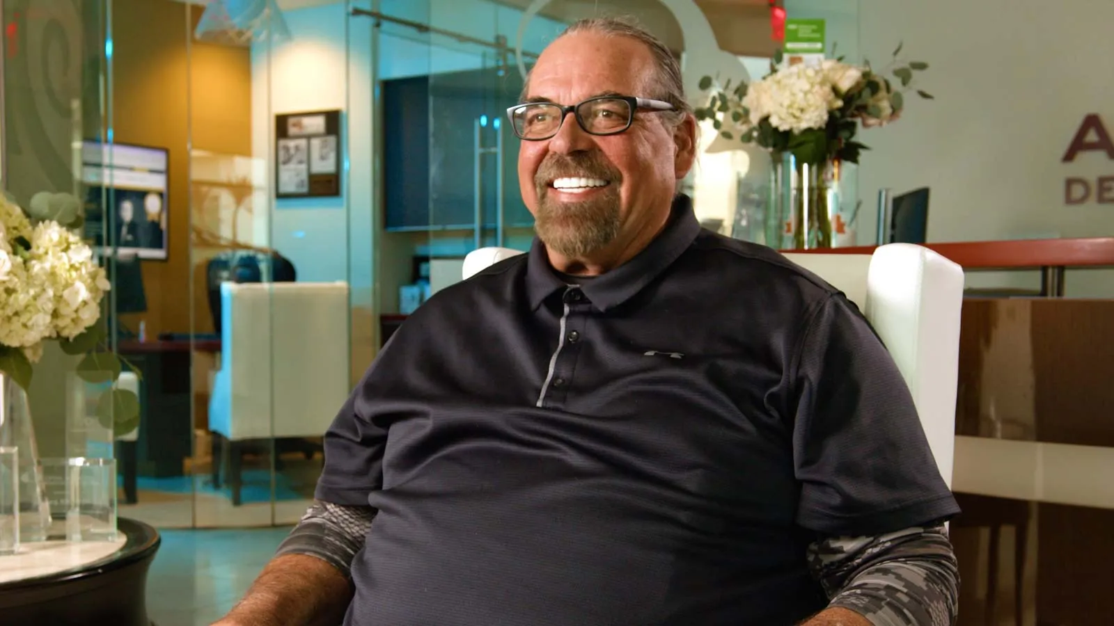 Smiling middle-aged man with glasses sitting in a modern office with flowers and glass walls.