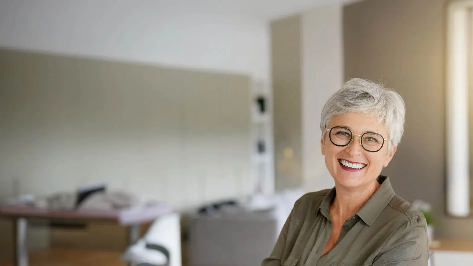 Smiling senior woman with short gray hair and glasses in a bright, modern living room.