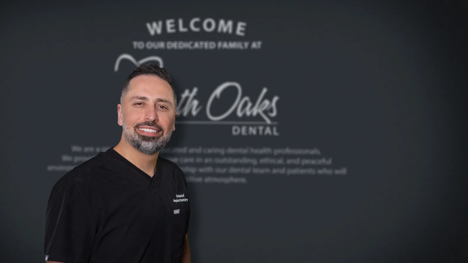 Smiling male dental professional in black scrubs stands in front of a North Oaks Dental welcome sign.