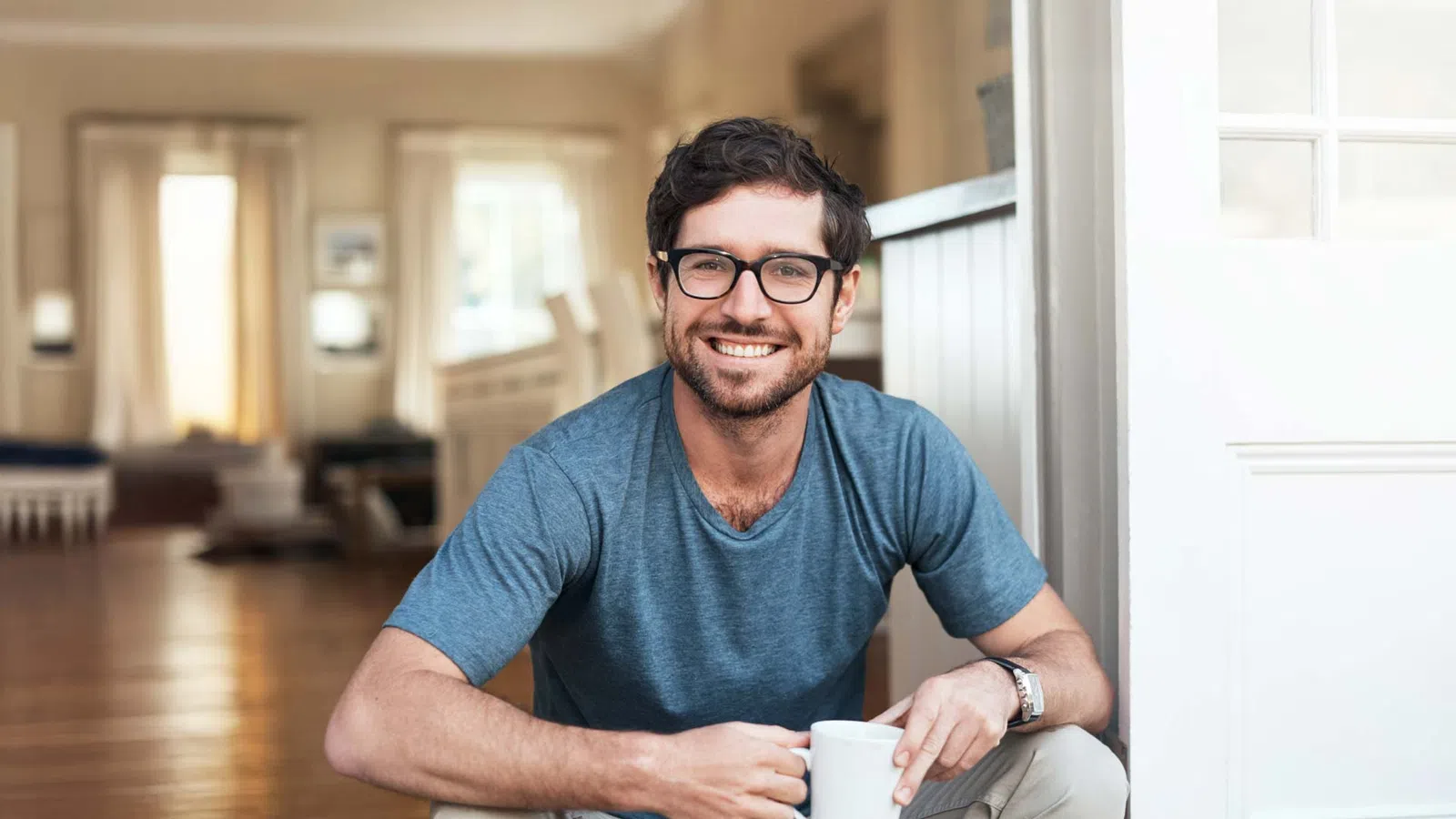 Smiling man wearing glasses and a blue shirt holding a white mug sitting indoors by a door.