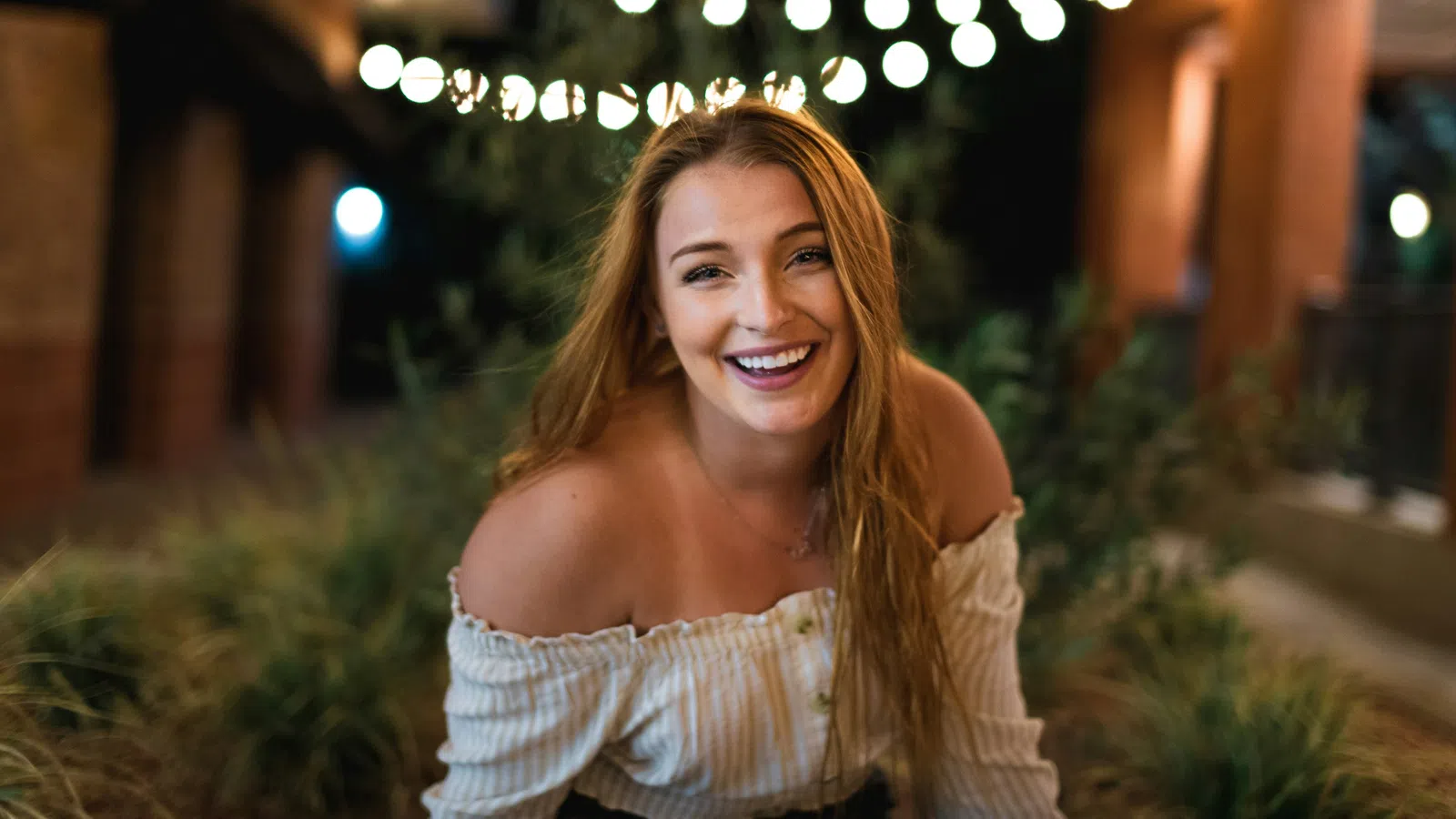 Smiling young woman with long hair in white off-shoulder top under string lights at night outdoors.