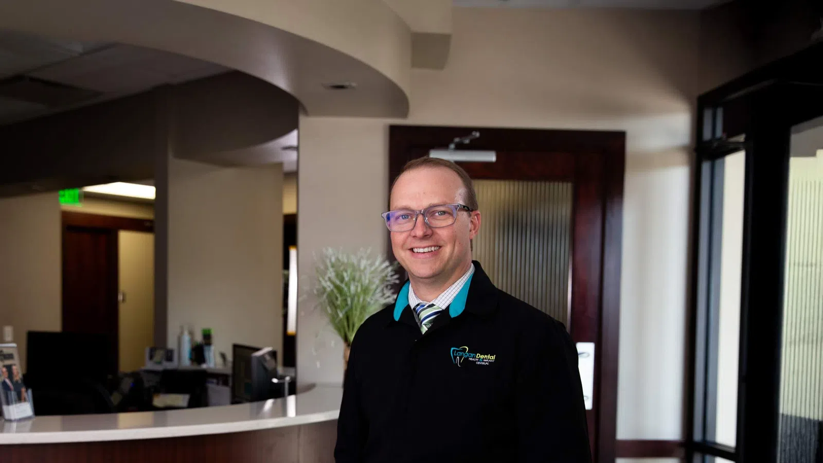 Smiling male dentist wearing glasses and uniform standing in modern dental office reception area.