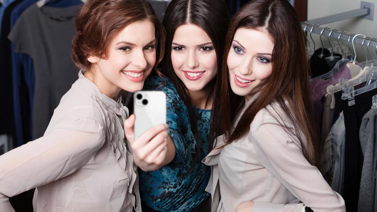 Three young women smiling and taking a selfie together in a clothing store with racks of clothes in the background