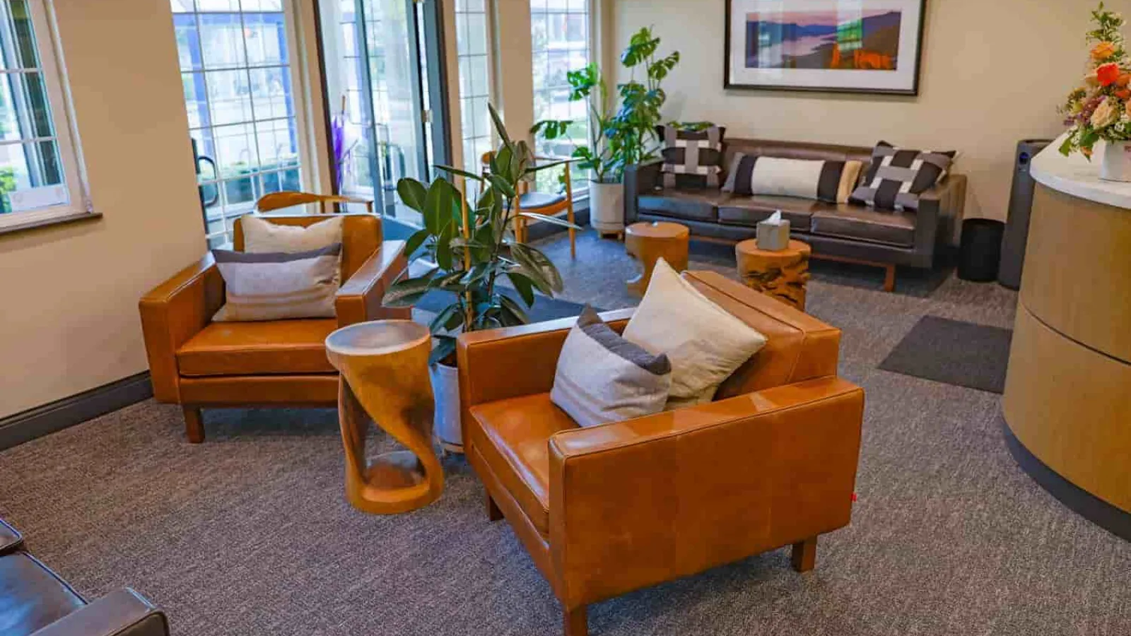 Modern living room with brown leather chairs, plants, gray sofa, wooden tables, and framed artwork on the wall
