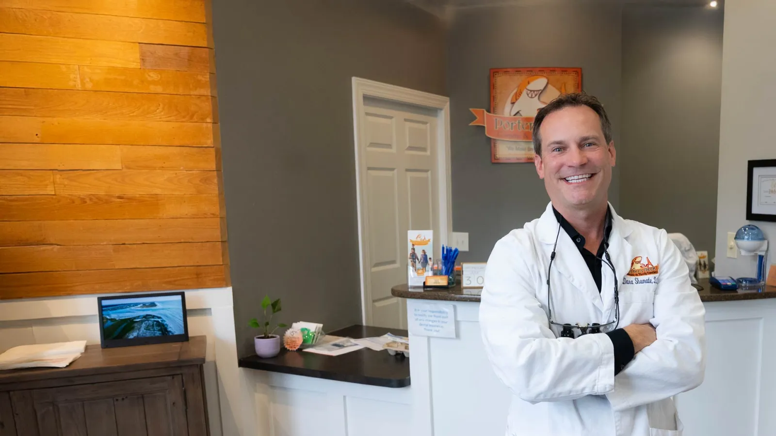 Smiling male dentist in white coat stands with arms crossed in modern dental office reception area.