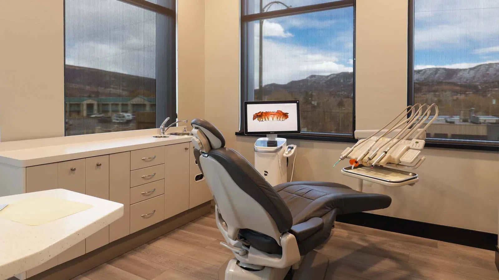 Modern dental exam room with reclining chair, dental tools, computer screen, and large windows showing mountainous view.