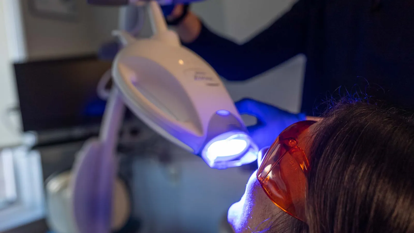 Person wearing protective glasses receiving dental teeth whitening treatment with blue light device.