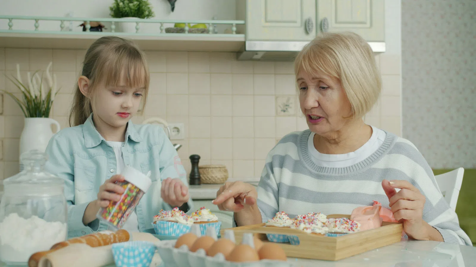 Grandmother and granddaughter decorating cupcakes together in a bright kitchen with baking ingredients on the table