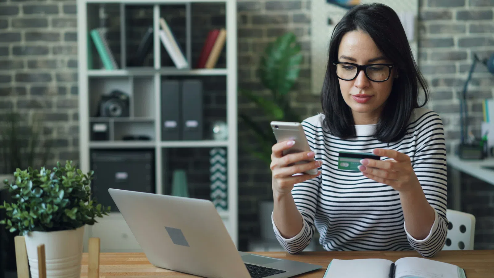 Woman wearing glasses using smartphone and credit card for online shopping at a desk with laptop and notebook.