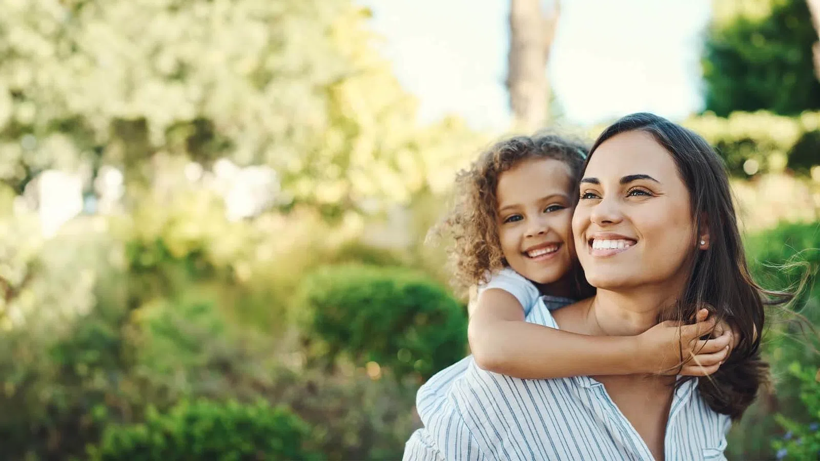 Smiling mother giving her daughter a piggyback ride outdoors in a sunny green park.