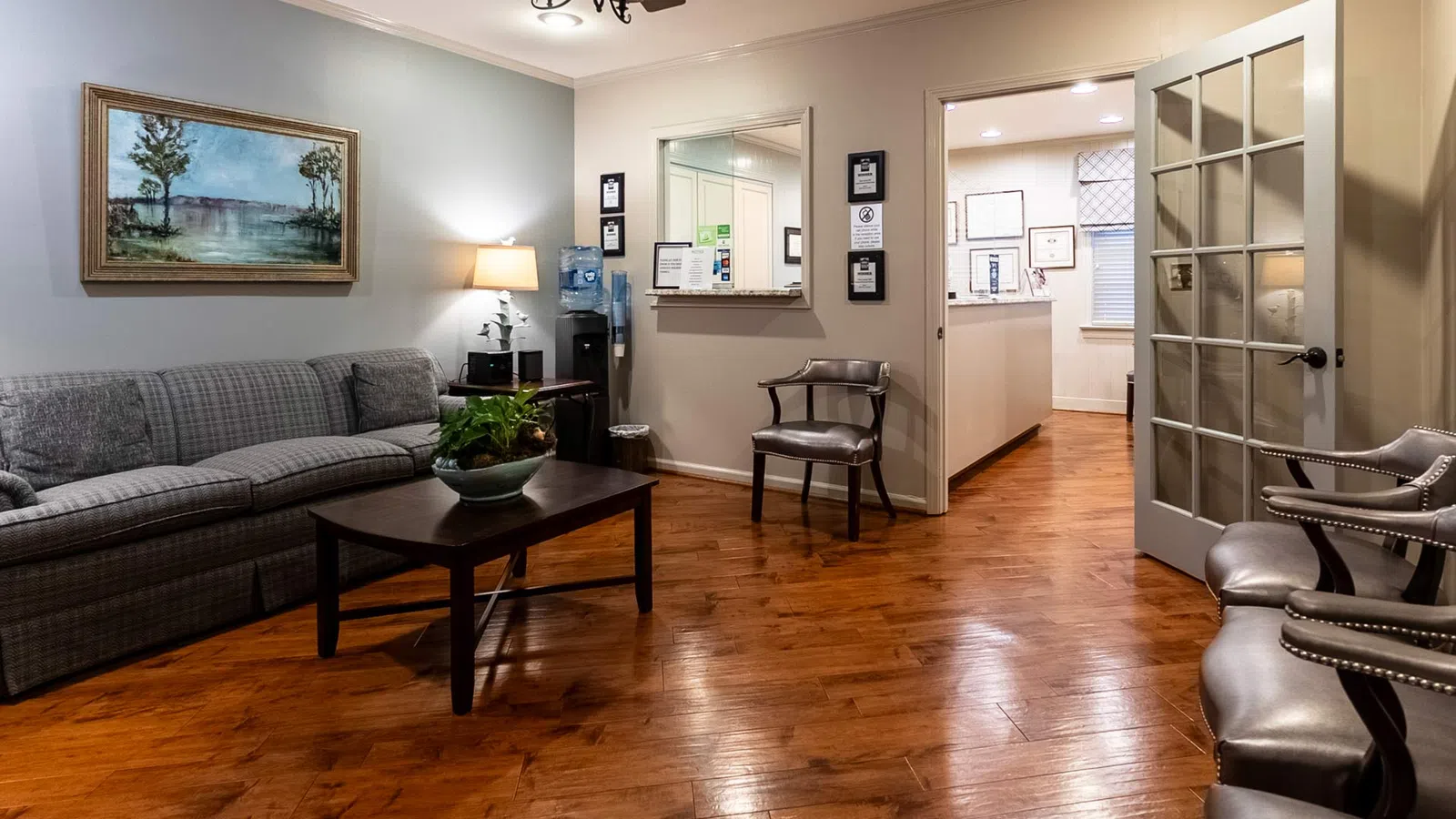 Cozy waiting room with wooden floors, gray sofa, chairs, coffee table, wall art, and a water dispenser.