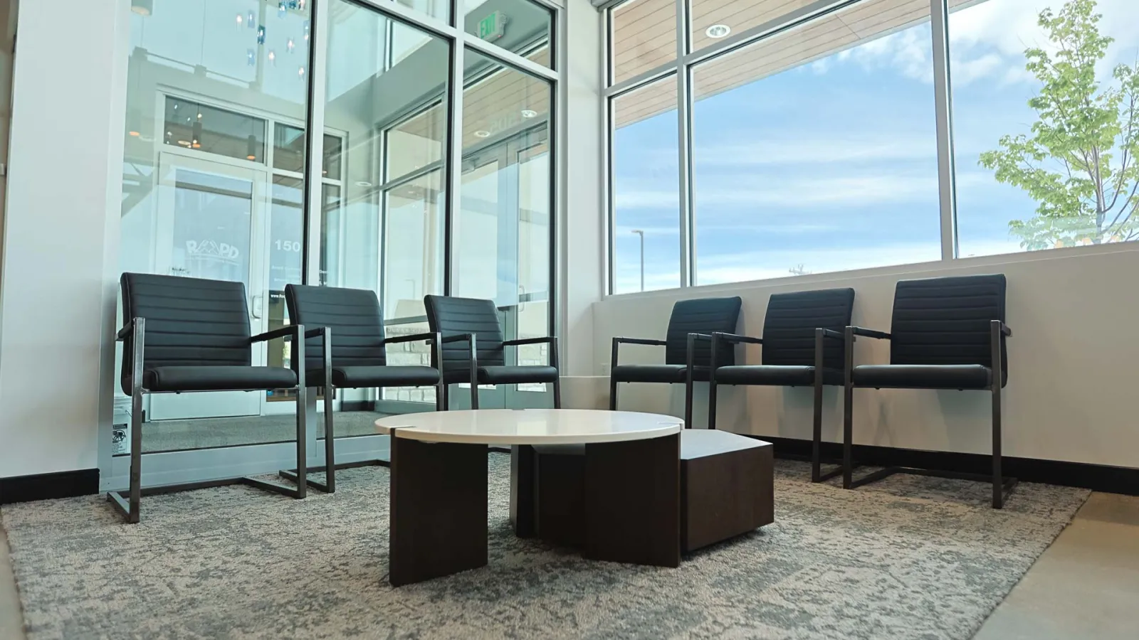 Modern waiting area with black chairs around a low round table on a gray carpet near large windows.