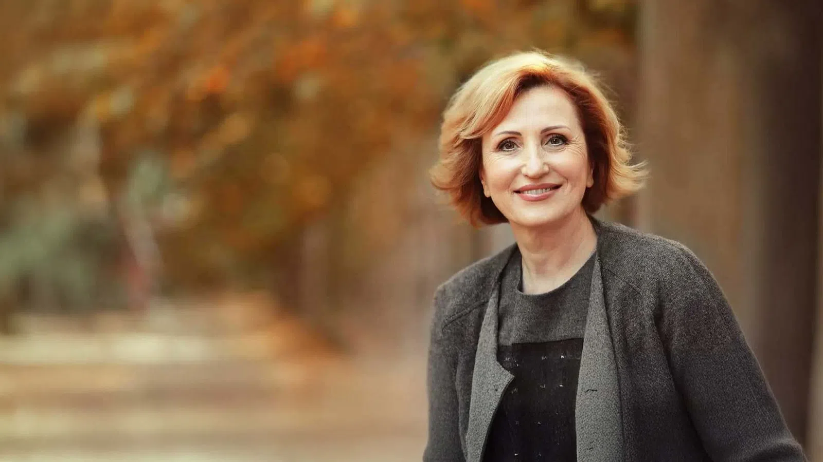 Smiling middle-aged woman with short blonde hair wearing a dark coat outdoors in autumn setting
