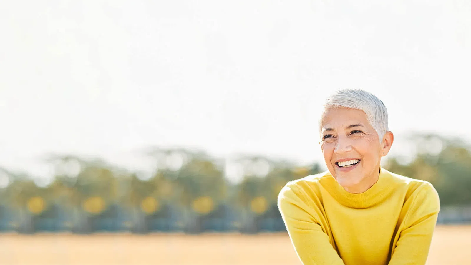 Smiling mature woman with short white hair wearing a bright yellow sweater outdoors with blurred trees background