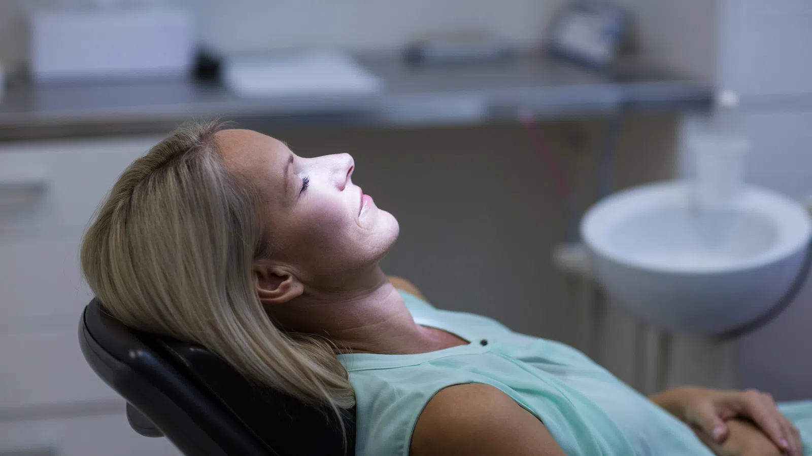 Woman reclining in dental chair with eyes closed, illuminated by bright dental light in clinic