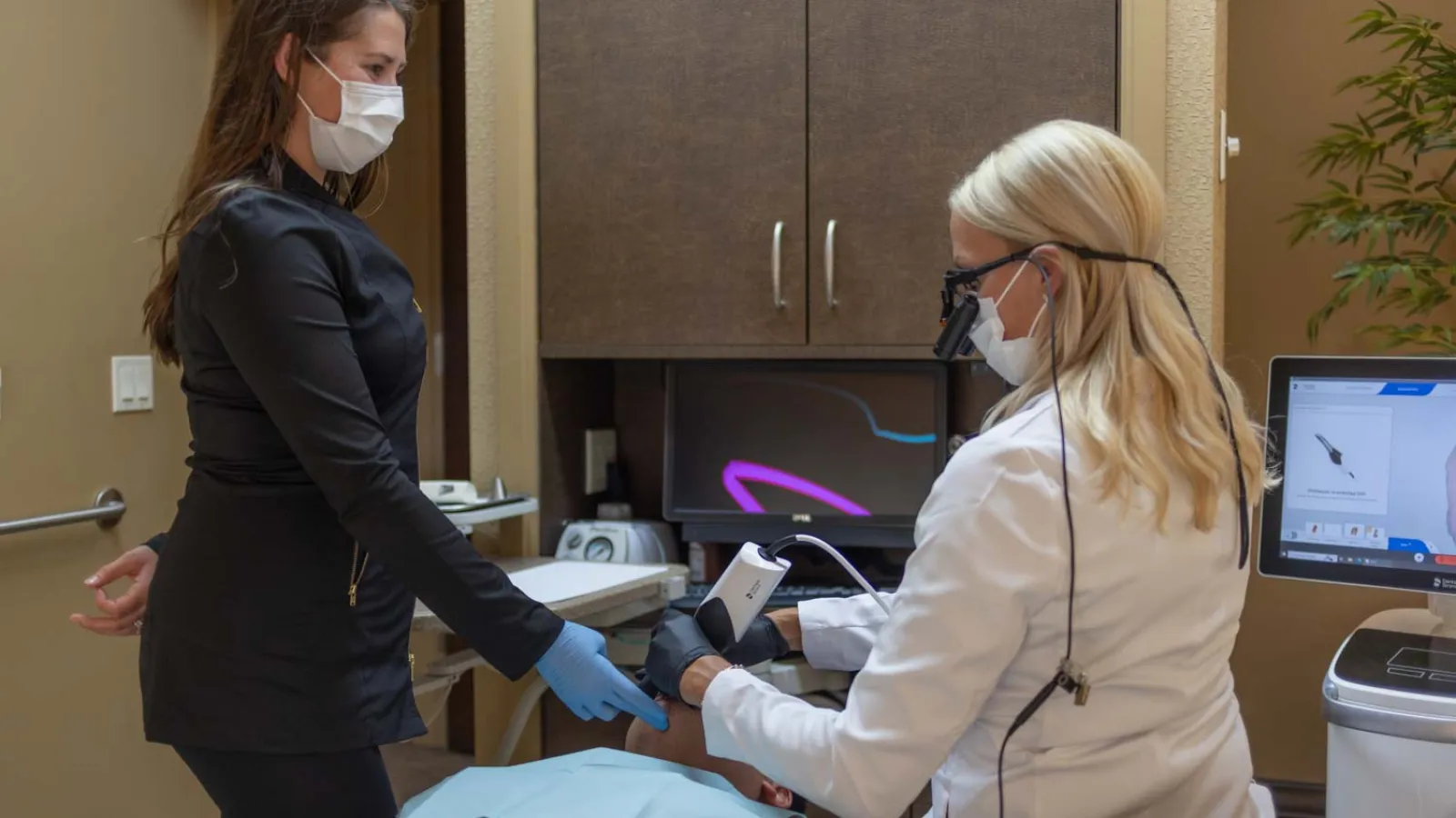Dentist using digital scanner on patient's teeth while assistant supports patient in clinical room with masks.