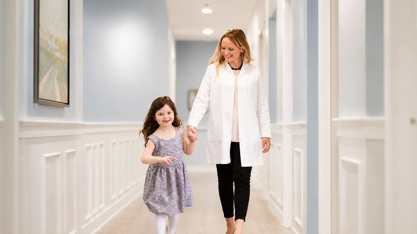 Female doctor holding hands with smiling young girl walking down a bright hallway in a medical clinic.