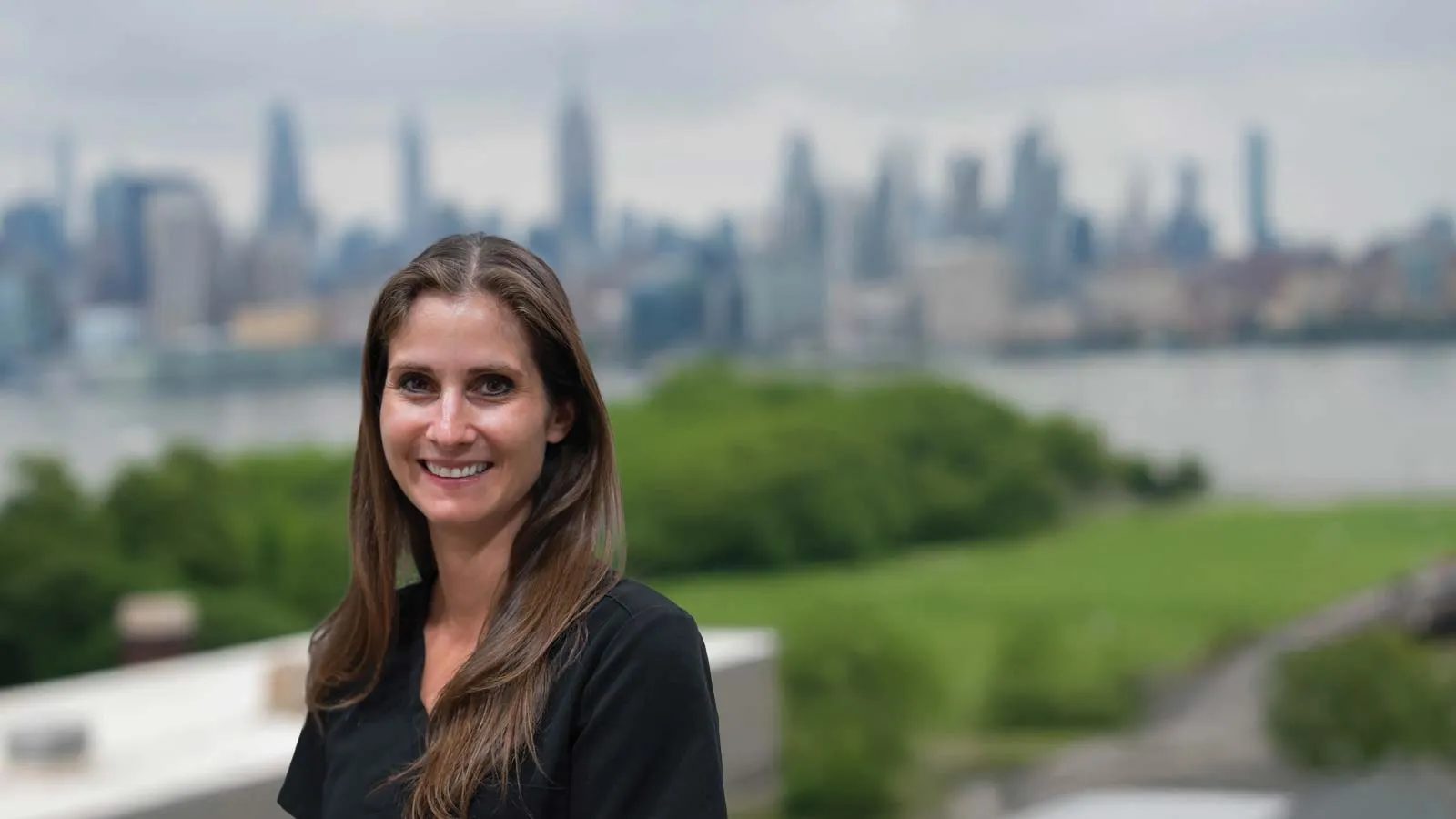 Smiling woman with long brown hair standing outdoors with greenery and city skyline in the background.