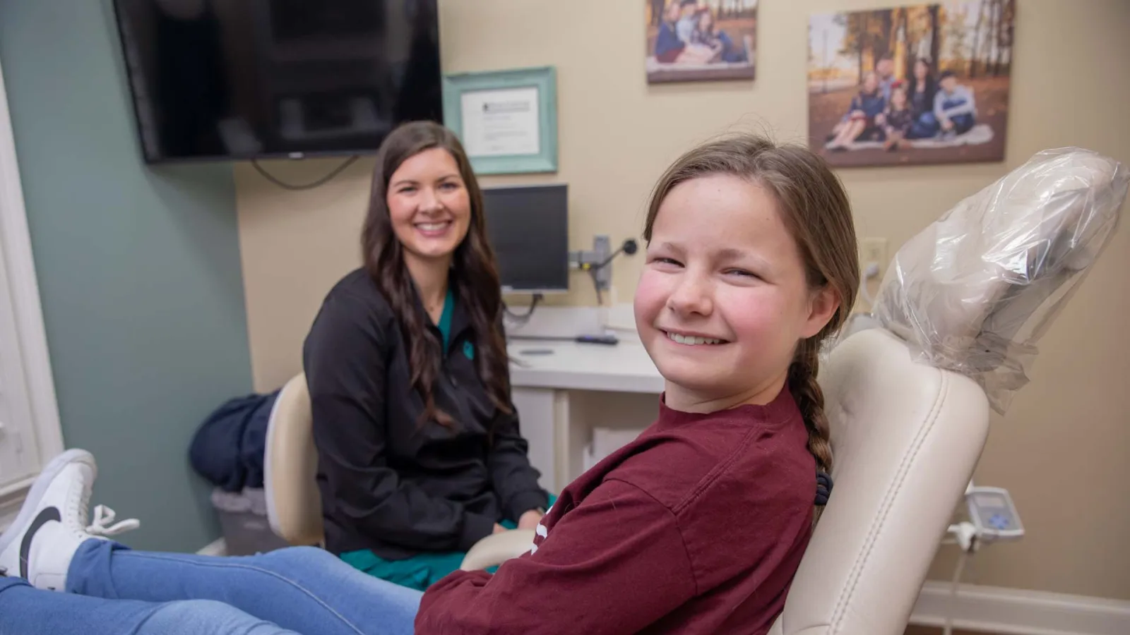Smiling young girl sitting in dental chair with female dentist in background in bright dental office.