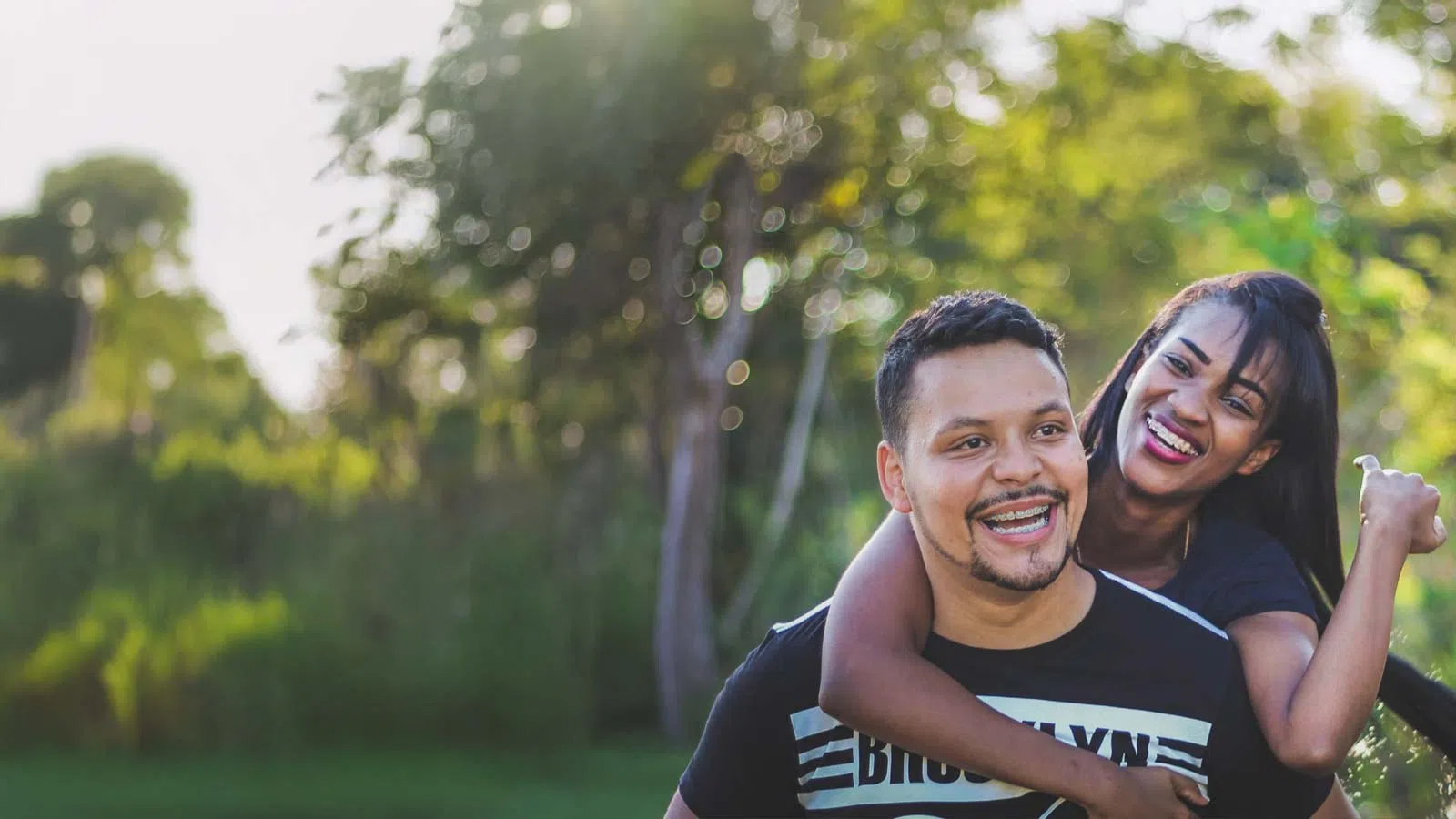 Happy young couple enjoying piggyback ride outdoors in lush green park with bright sunlight.