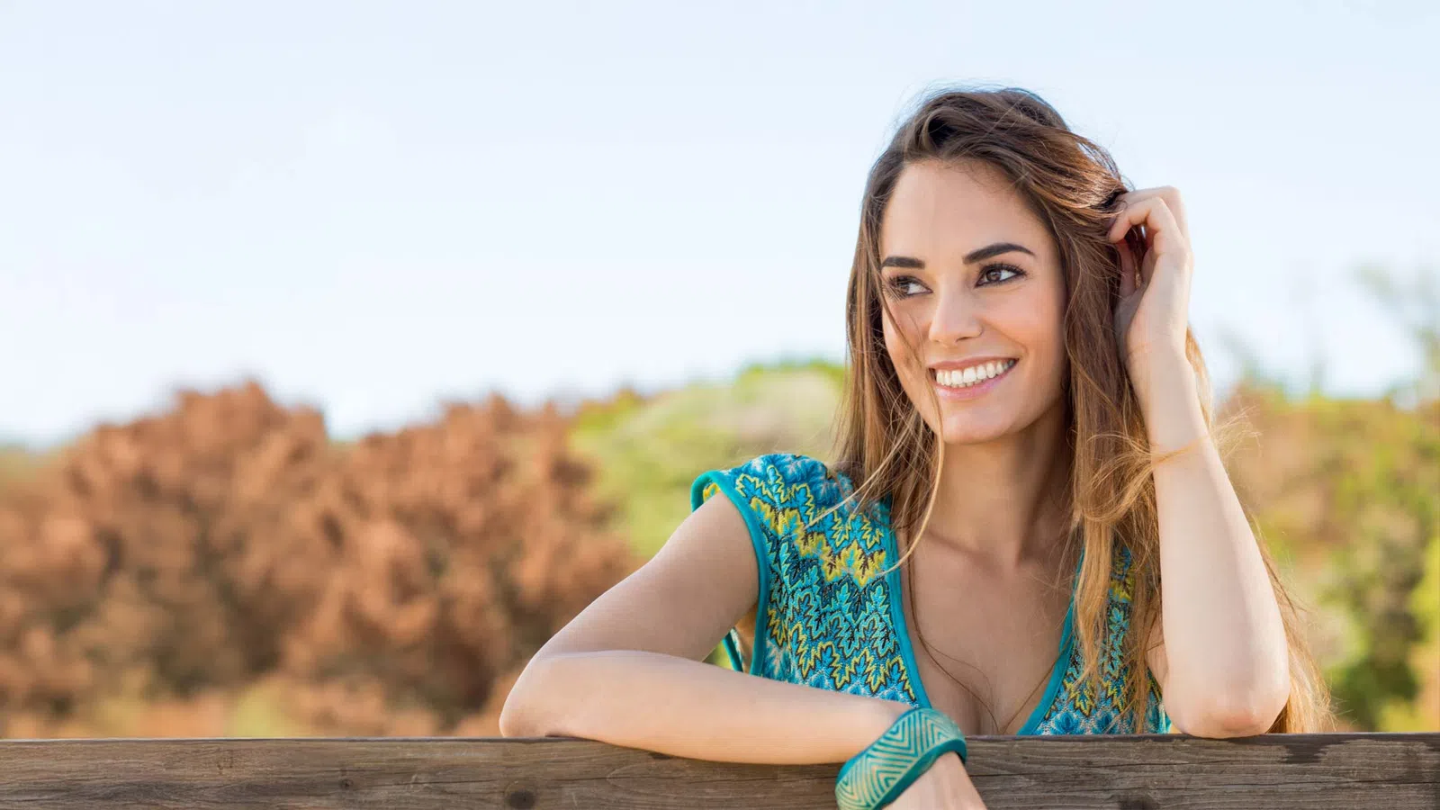 Young woman smiling outdoors leaning on a wooden fence with autumn trees in the background