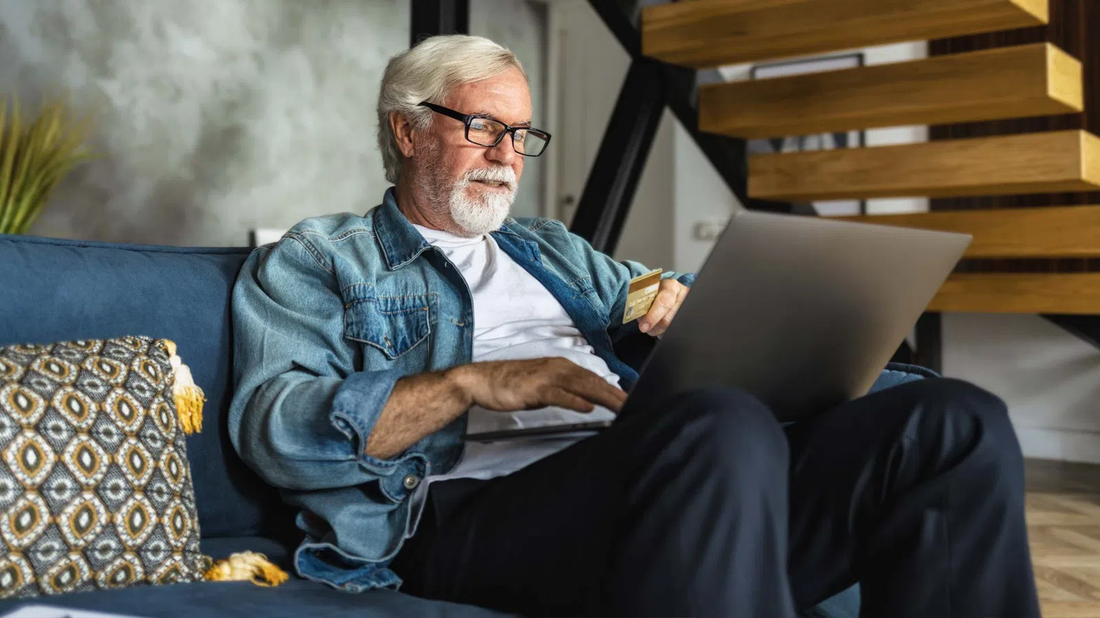Senior man with glasses sitting on couch using laptop to shop online holding credit card.