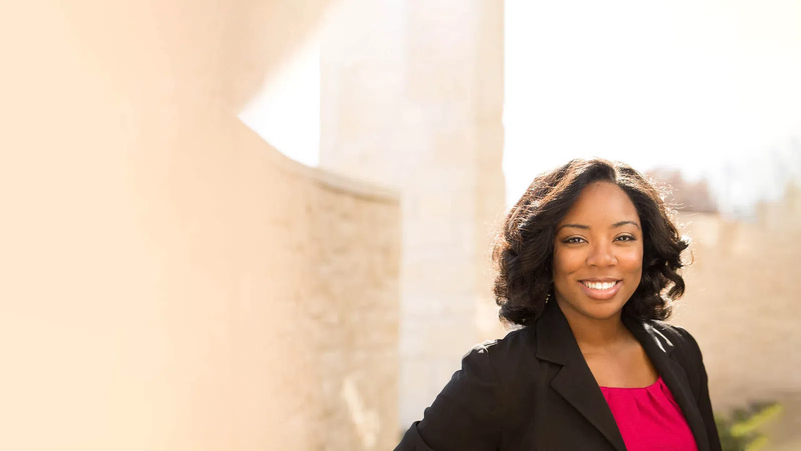 Confident woman in business attire smiling outdoors with soft sunlight and stone wall background