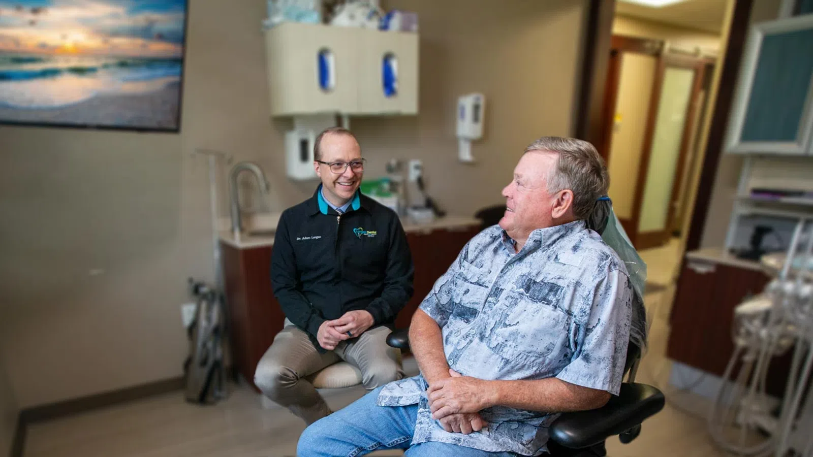 A dentist consulting with a patient in a modern dental office with calming decor and beach-themed artwork.