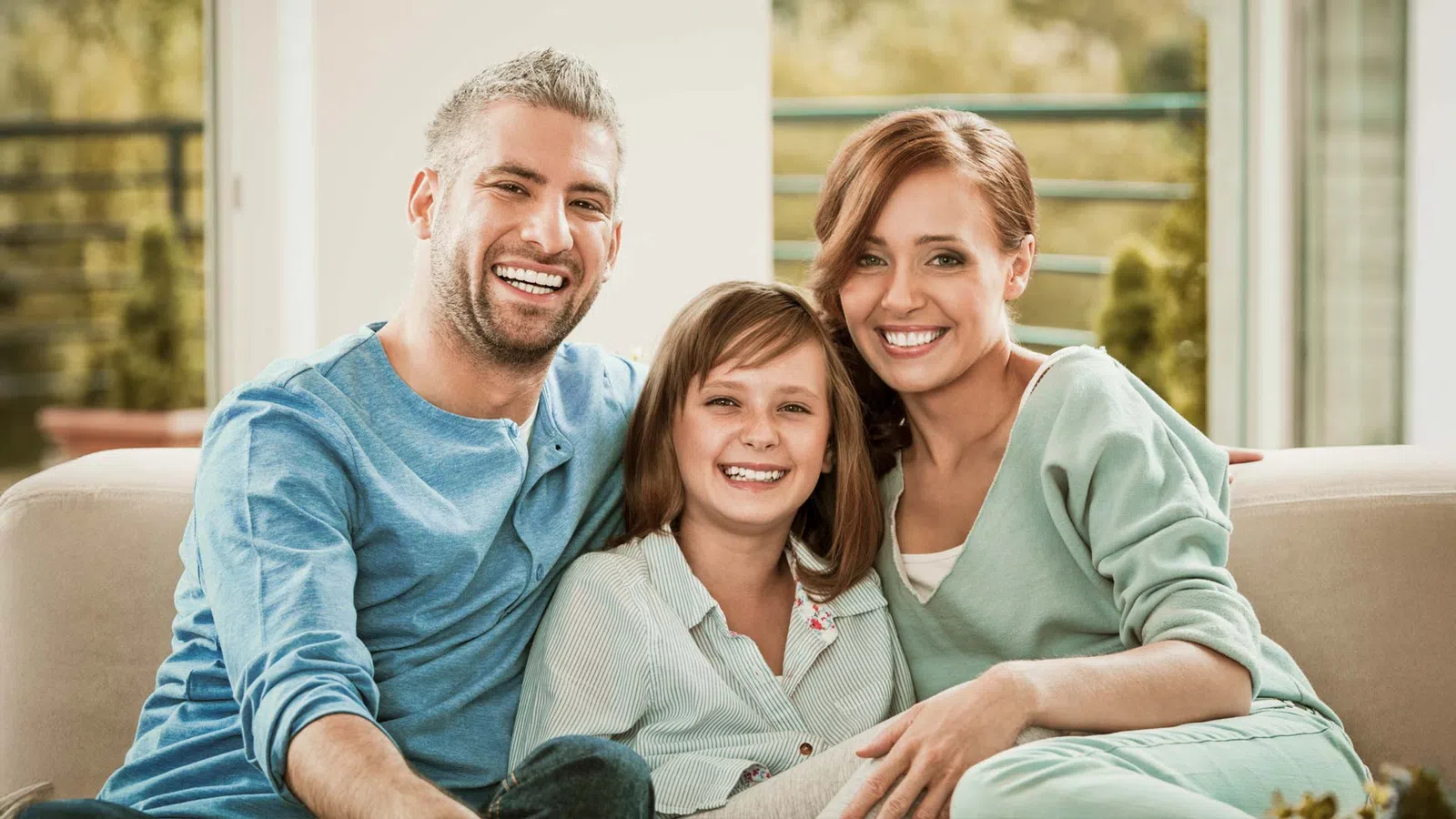 Smiling family of three sitting closely on a couch in a bright living room, showing warmth and happiness.