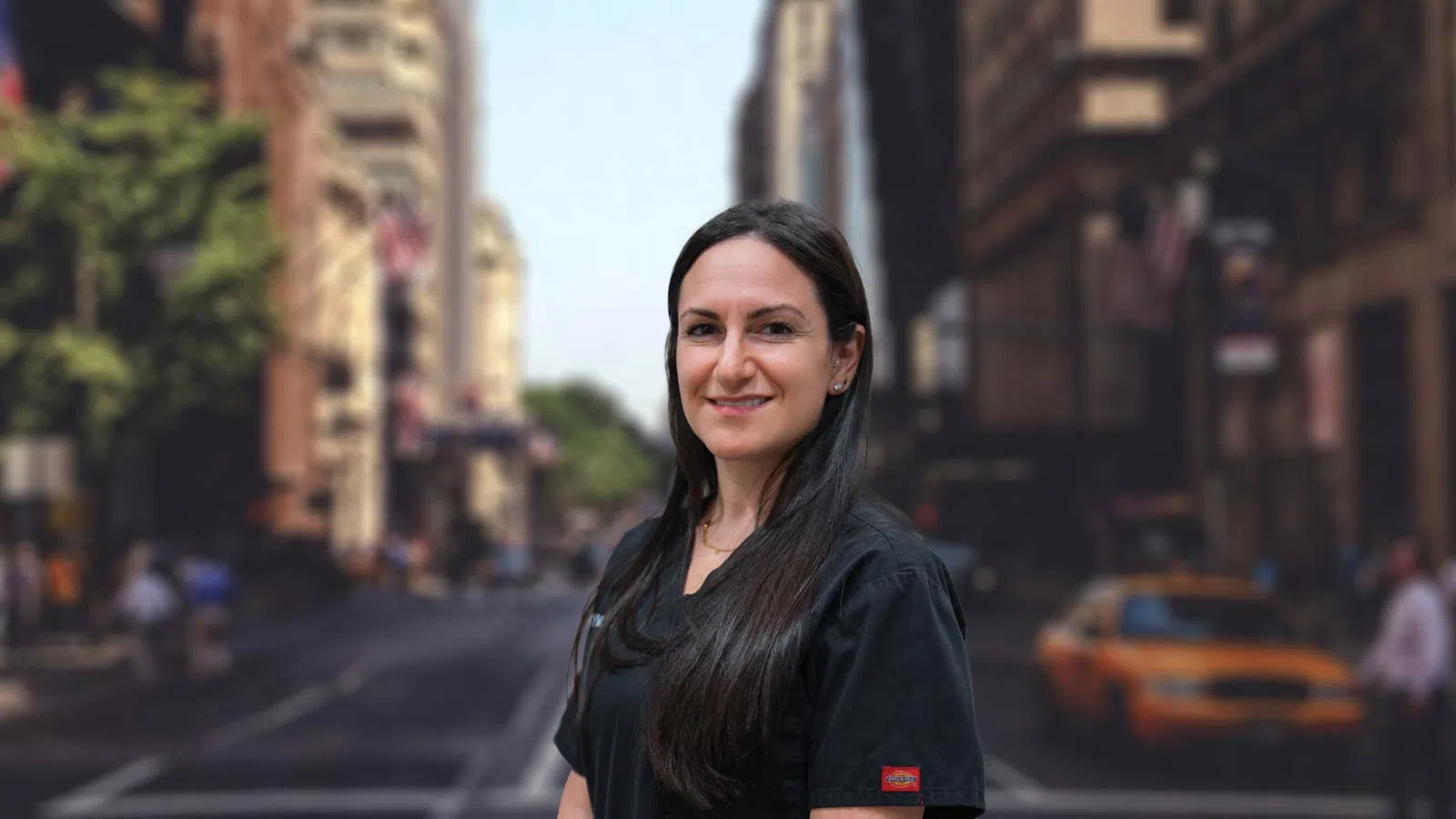 Smiling woman with long dark hair in black scrubs stands on a city street with blurred buildings and cars.