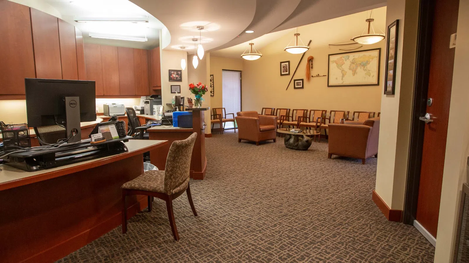 Spacious waiting area with wooden reception desk, computer, comfortable chairs, and wall decorations in warm lighting.