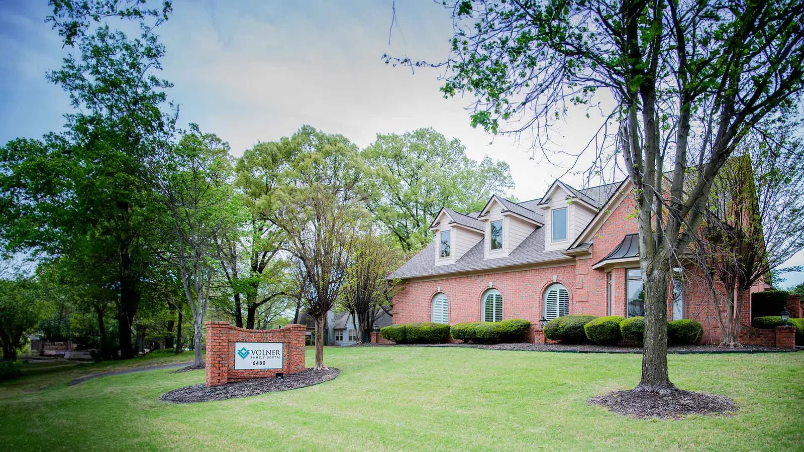 Brick office building with green lawn and trees under a cloudy blue sky during springtime.