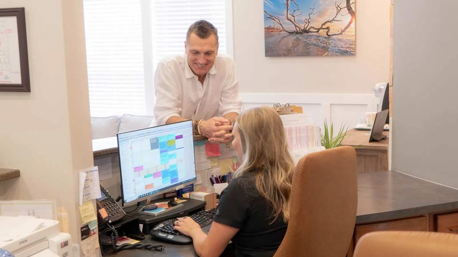 Man and woman discussing work at a computer desk showing a colorful calendar on screen in bright office