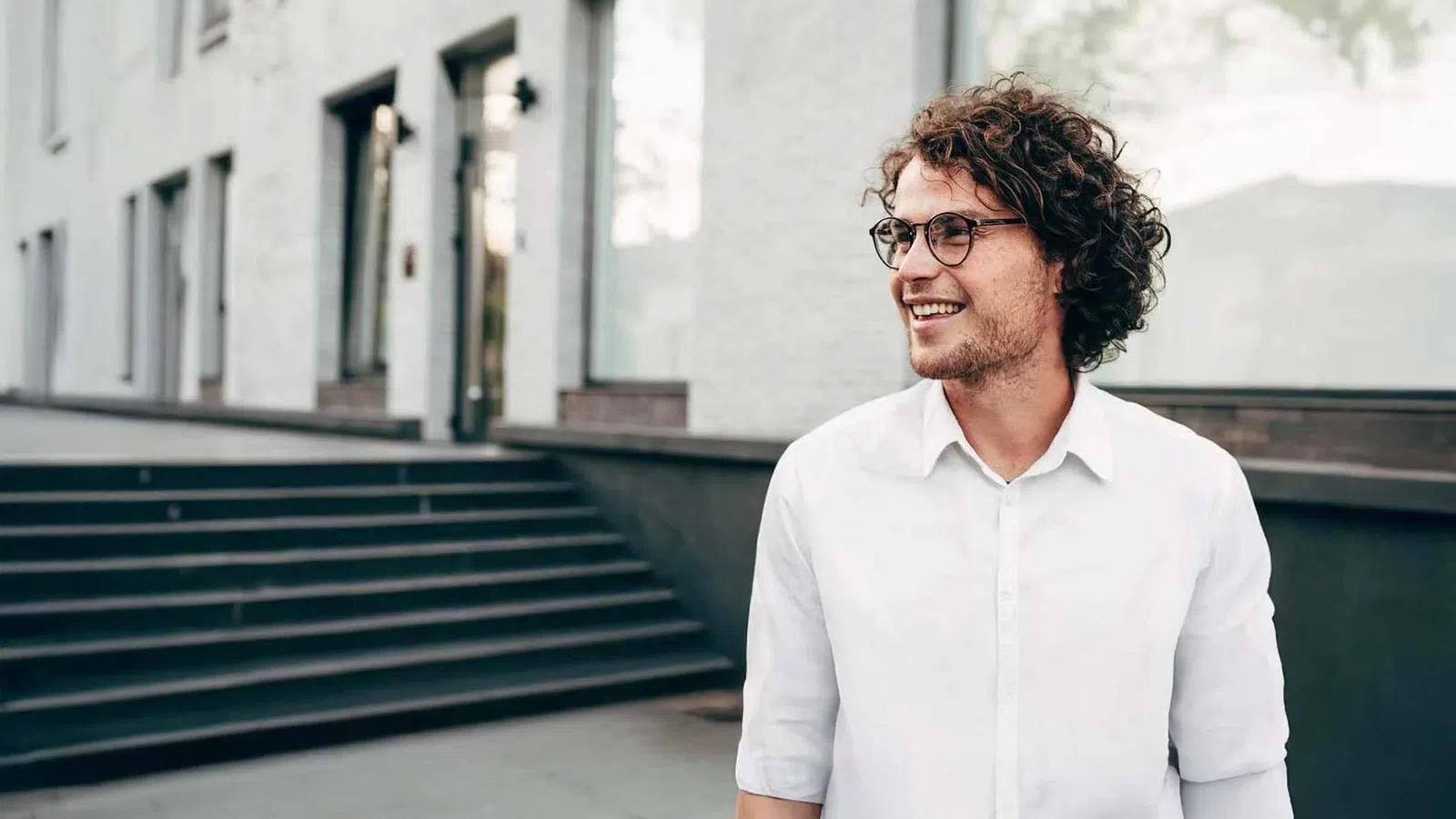 Young man with curly hair and glasses wearing white shirt smiling outdoors near modern building steps.