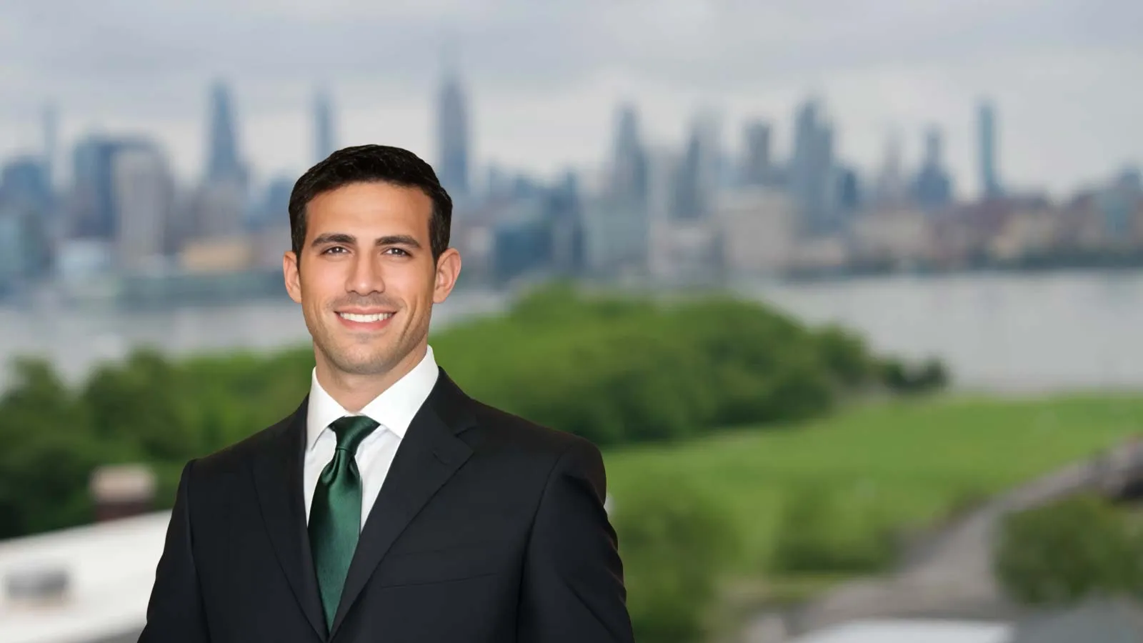 Young businessman in dark suit and green tie smiling with blurred city skyline and greenery in the background.