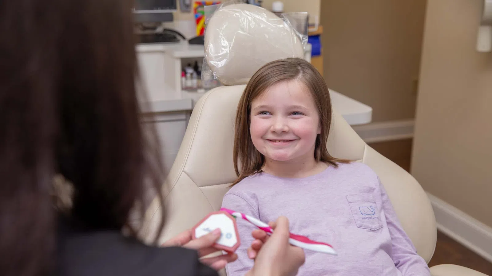 Smiling young girl in dental chair learning toothbrushing techniques from dental professional using model.