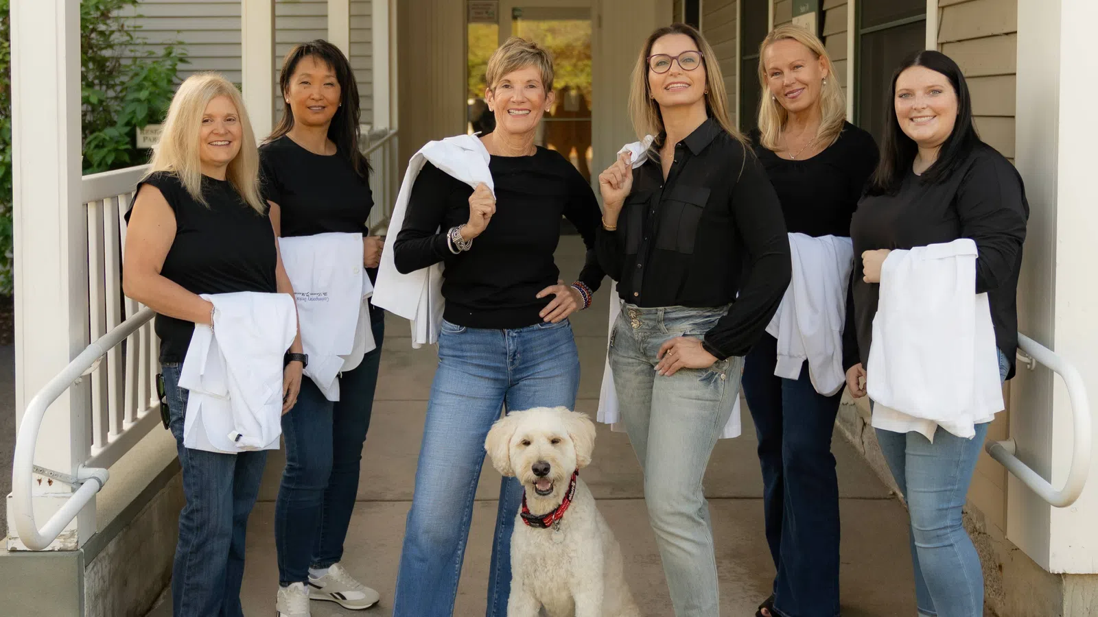 Six women in black tops and jeans holding white shirts on a porch with a smiling white dog sitting in front.