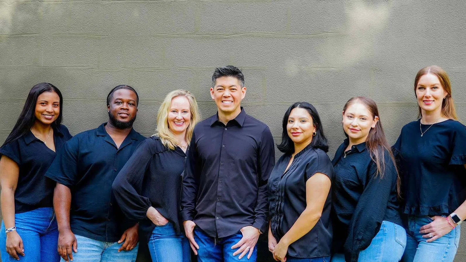 Diverse group of seven young professionals standing against a gray wall wearing black tops and blue jeans.