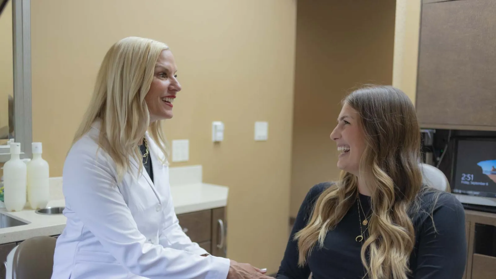 Female doctor and patient smiling and holding hands in a medical office consultation setting.