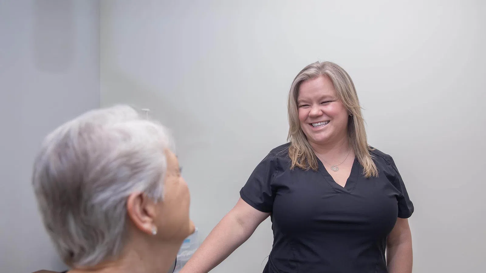 Smiling healthcare professional in black scrubs talking to an elderly patient in a clinic setting.