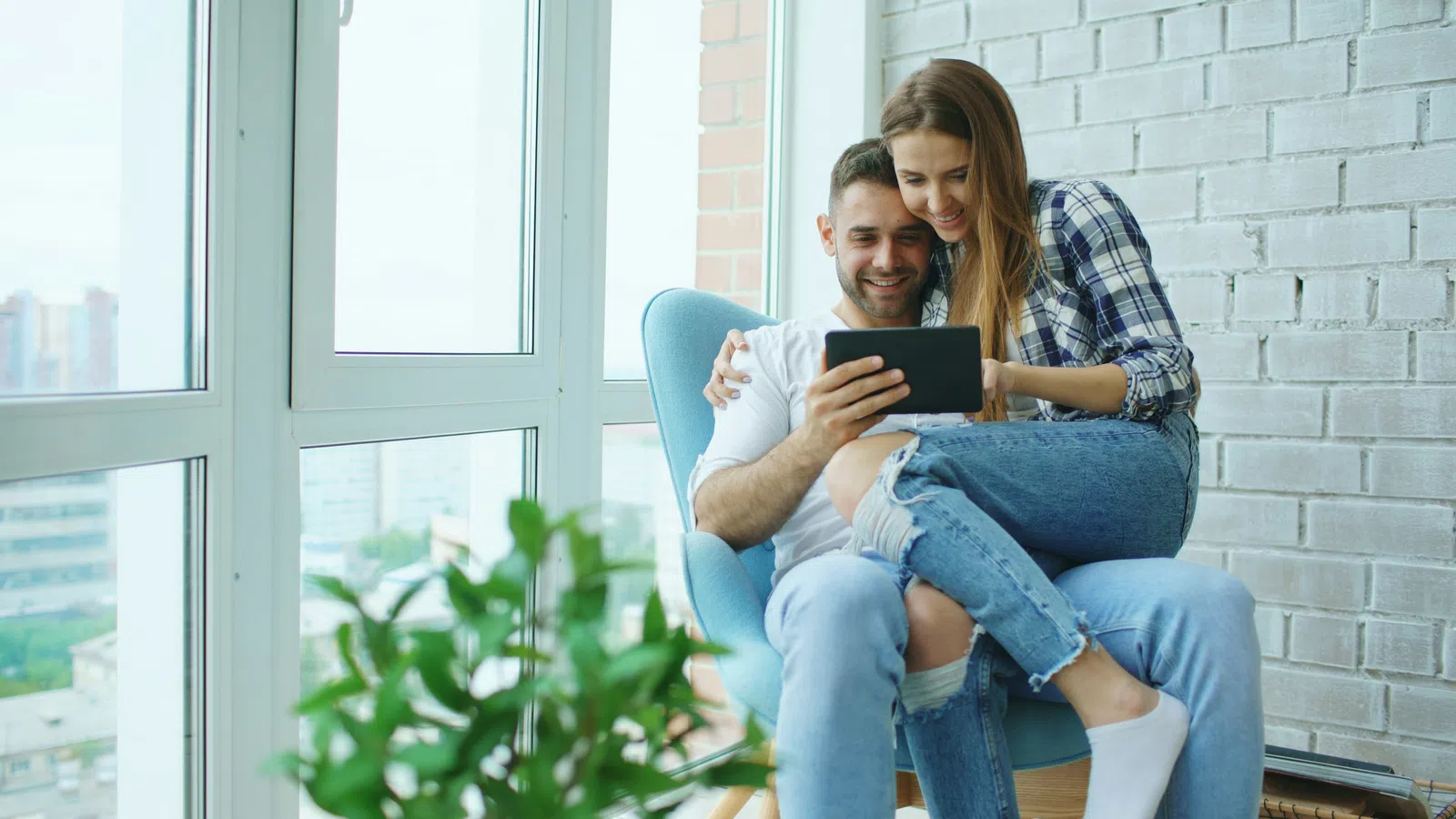 Young couple sitting on a chair by a window, smiling and looking at a tablet together indoors.