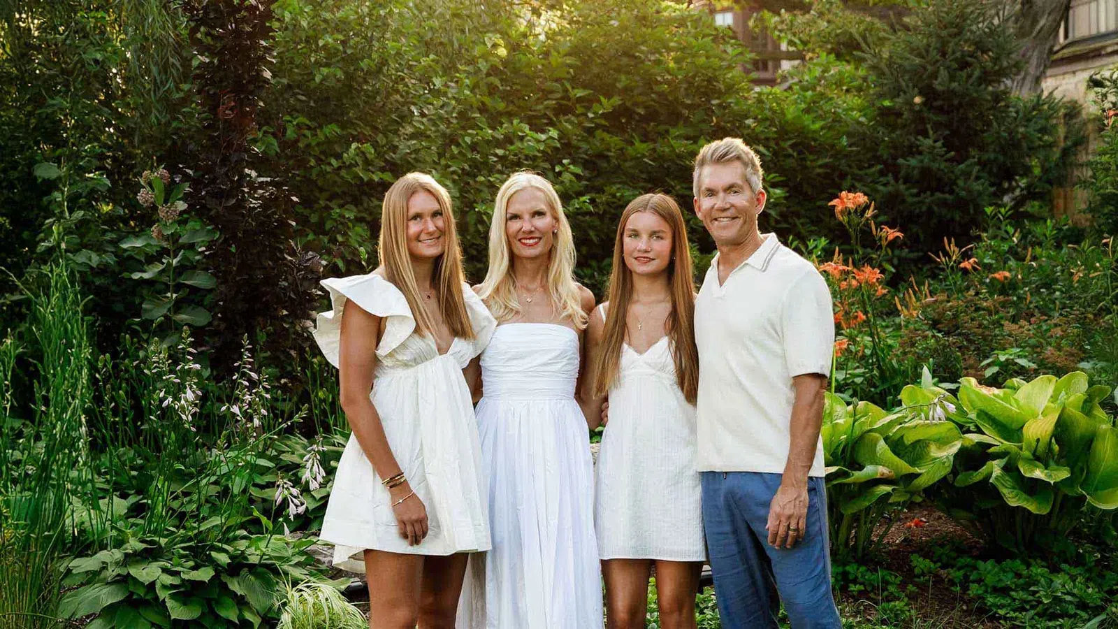 Happy family of four dressed in white standing together in a lush garden with greenery and flowers.