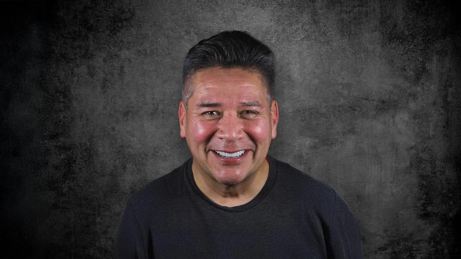 Smiling man with dark hair wearing a black shirt against a textured dark gray background