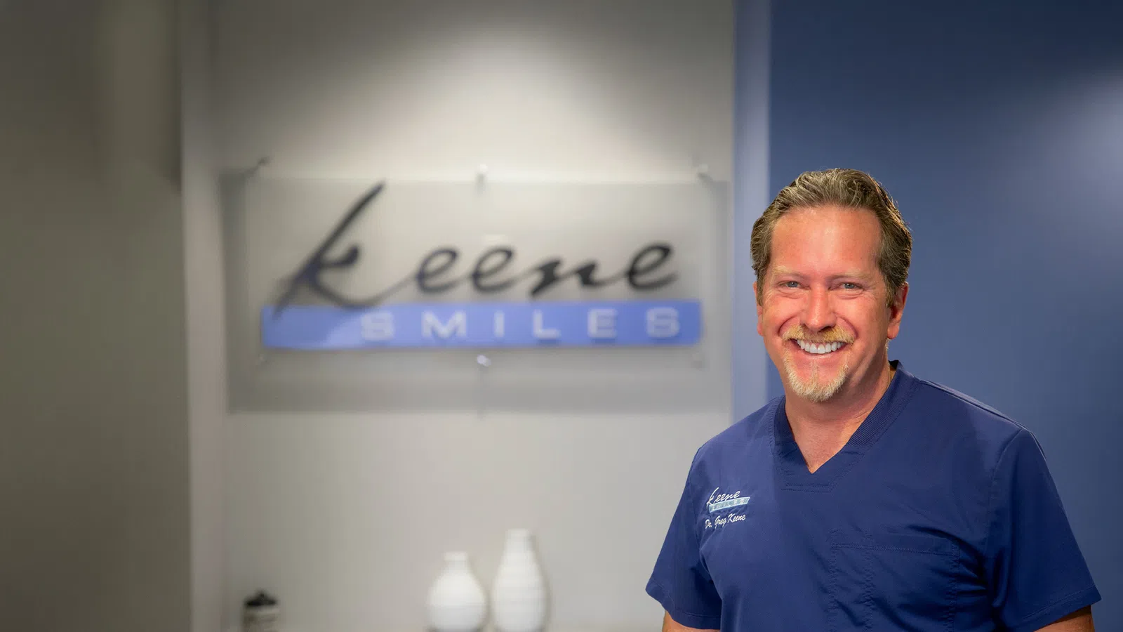 Smiling male dentist in blue scrubs standing in front of Keene Smiles dental office sign.