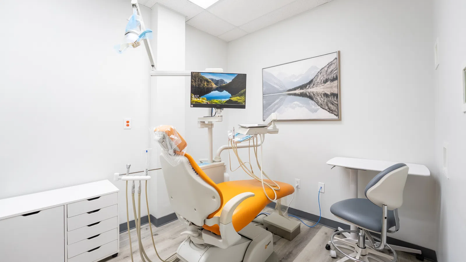 Modern dental office with orange patient chair, dental equipment, monitor, and nature-themed wall art in white room.