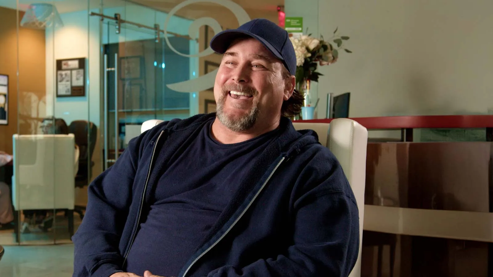 Smiling man in navy blue hoodie and cap sitting relaxed in a modern office lounge with glass walls and furniture.