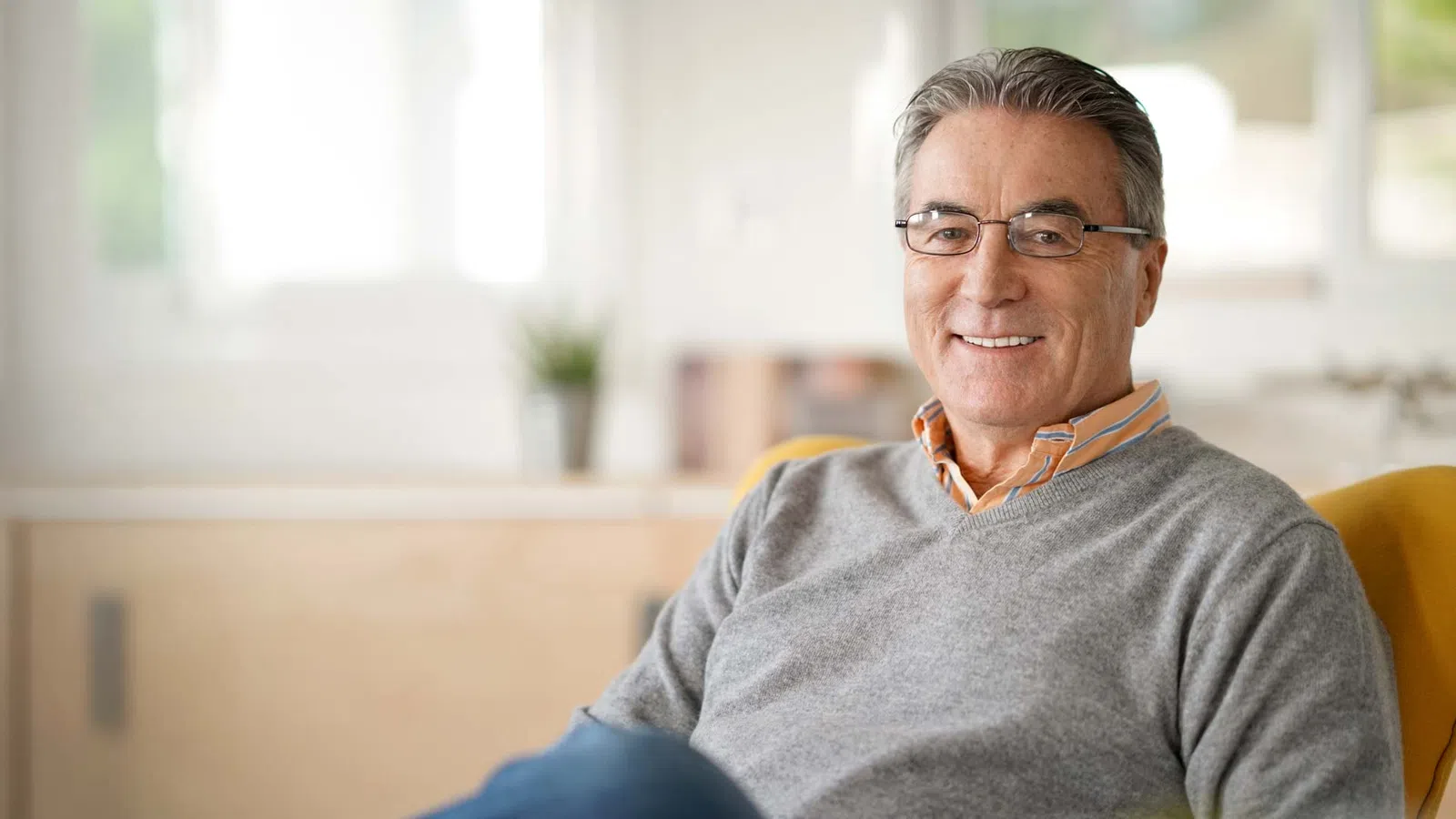 Smiling mature man wearing glasses and gray sweater sitting indoors with blurred background.