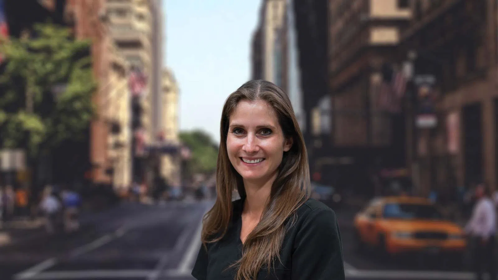 Smiling woman with long brown hair in black top stands in a blurred urban city street with buildings and yellow taxi.